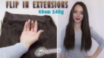 Ženska z dolgimi ravnimi rjavimi lasmi stoji nasmejana z rokami na bokih. Na njeni levi je slika prikazana z roko, ki drži lasni podaljšek z besedilom nad in pod njim »FLIP IN EXTENSIONS« in »40 cm 140 g.