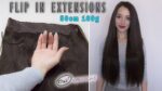 Roka, ki prikazuje komplet rjavih lasnih podaljškov na belem ozadju z besedilom "FLIP IN EXTENSIONS 50 cm 160 g." Na desni se smehlja ženska z dolgimi ravnimi rjavimi lasmi, oblečena v sivo majico. Pod roko je logotip »Oxy Extensions«.