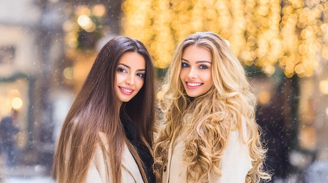 Dve ženski z dolgimi rjavimi in blond lasmi v prazničnem zimskem ambientu.